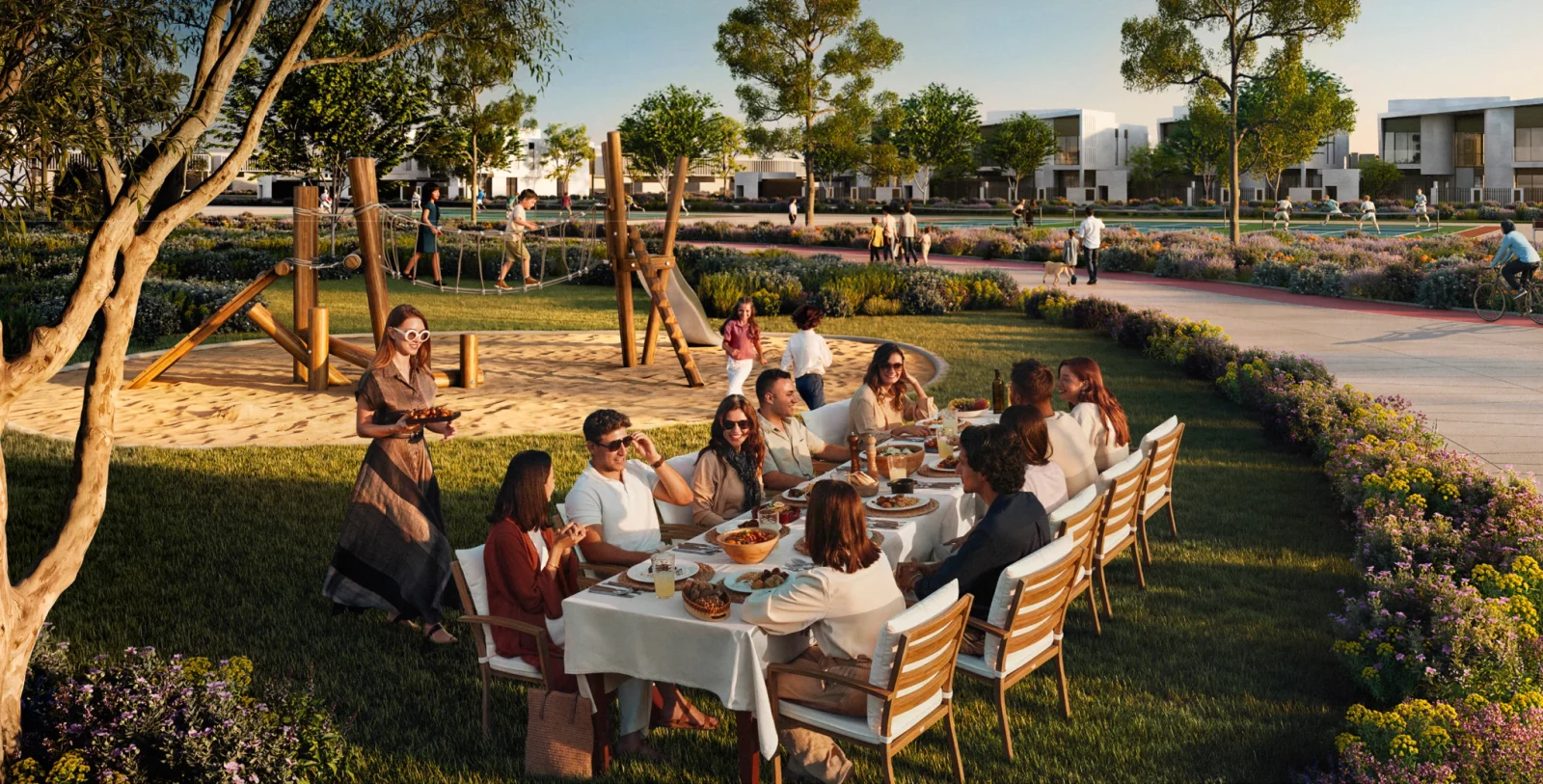 Render of a long dining table laid on a lawn in the Talay community park, with a rope-climbing playground, flowering planting, a cycling and jogging path, and rows of white flat-roofed houses behind
